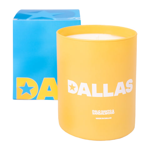 Yellow candle with white text "DALLAS PALO SANTO & SANDALWOOD MAD IN DALLAS" in front of blue box