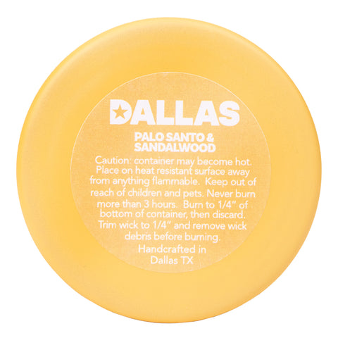 bottom side of yellow candle with white text "DALLAS PALO SANTO & SANDALWOOD Caution: container may become hot. Place on heat resistant surface away from anything flammable. Keep out of reach of children and pets. Never Burn more than 3 hours. Burn to 1/4" of bottom of container, then discard. Trim wick to 1/4" and remove wick debris before burning. Handcrafted in Dallas TX"